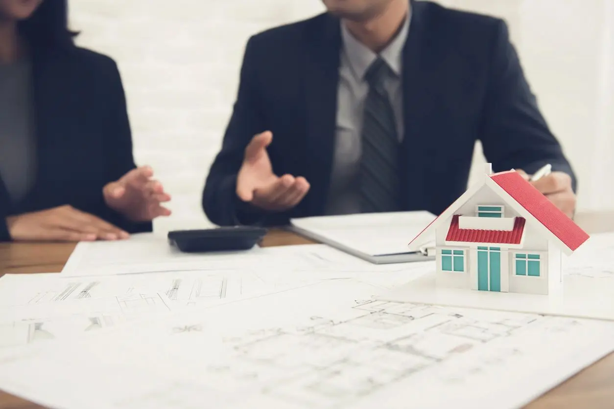 A man in a suit sitting at a table with papers and a model house.